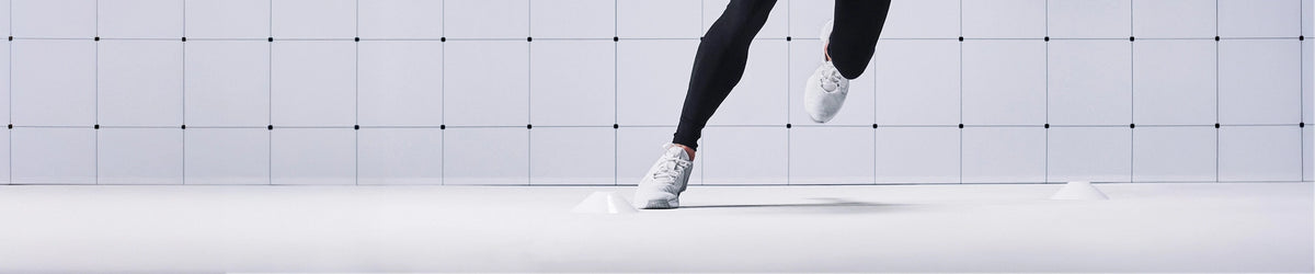 A person wearing black leggings and white athletic sneakers running against a minimalist white tiled wall and floor.