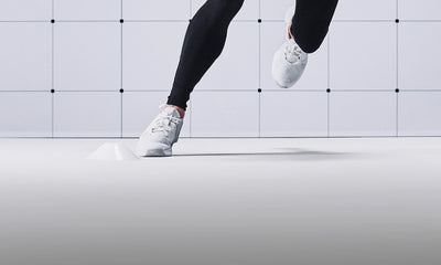 A person wearing black leggings and white athletic sneakers running against a minimalist white tiled wall and floor.