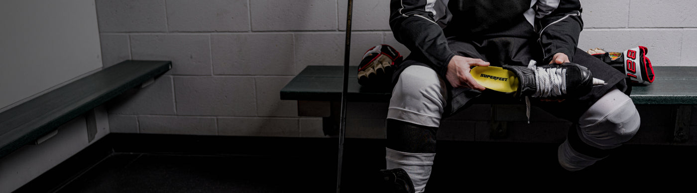 Person sitting on a bench in a hockey locker room with equipment. They are inserting a Superfeet Insole into their skates. 