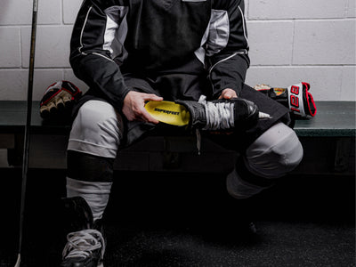 Person sitting on a bench in a hockey locker room with equipment. They are inserting a Superfeet Insole into their skates. 