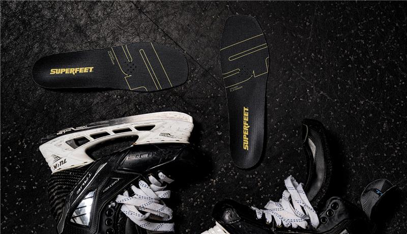Superfeet's Hockey Playmaker Insoles against a black background with Hockey equipment laying nearby. 