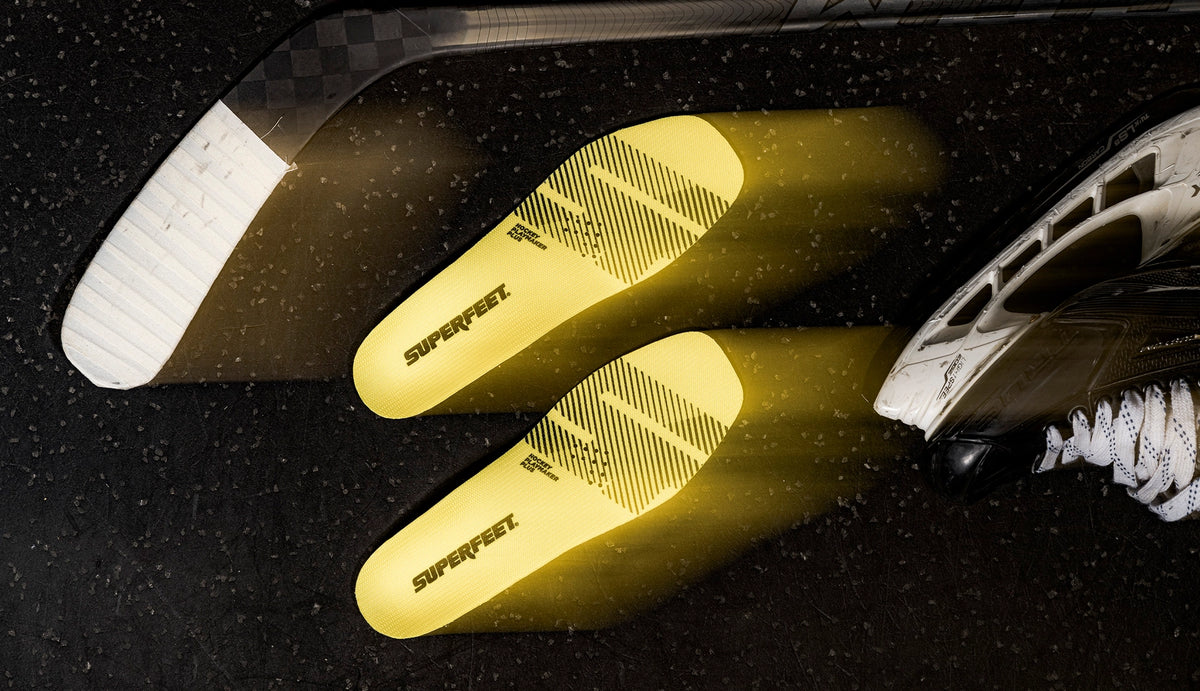 Superfeet's Hockey Playmaker Plus Insoles against a black background with Hockey equipment laying nearby. 